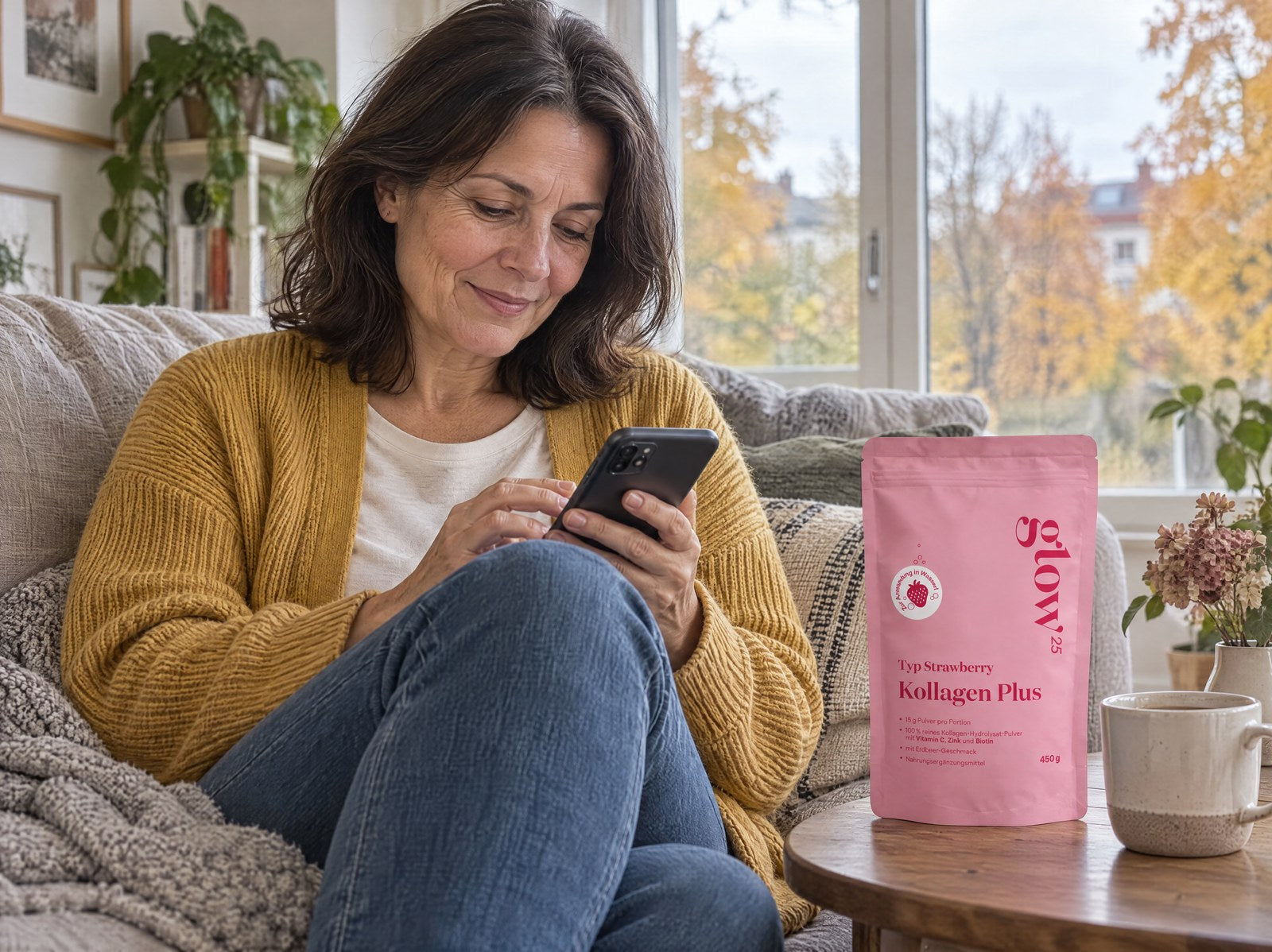 Frau im senffarbenen Cardigan wählt auf dem Sofa am Handy ihre nächste Lieferung, daneben Kollagen Plus Typ Strawberry