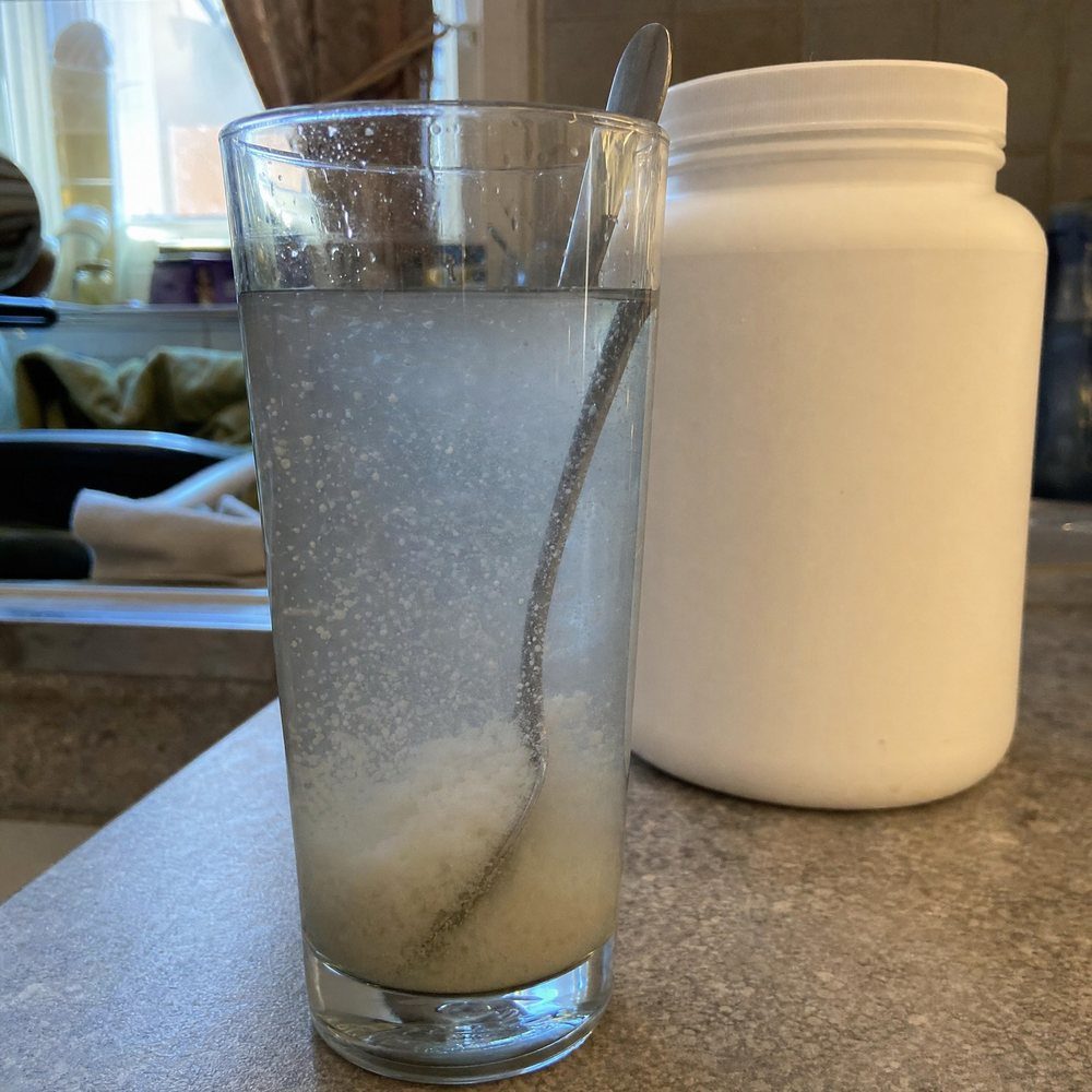 Un verre d'eau dans lequel une poudre de créatine bon marché ne se dissout pas et repose au fond comme du sable, derrière un pot blanc sans nom