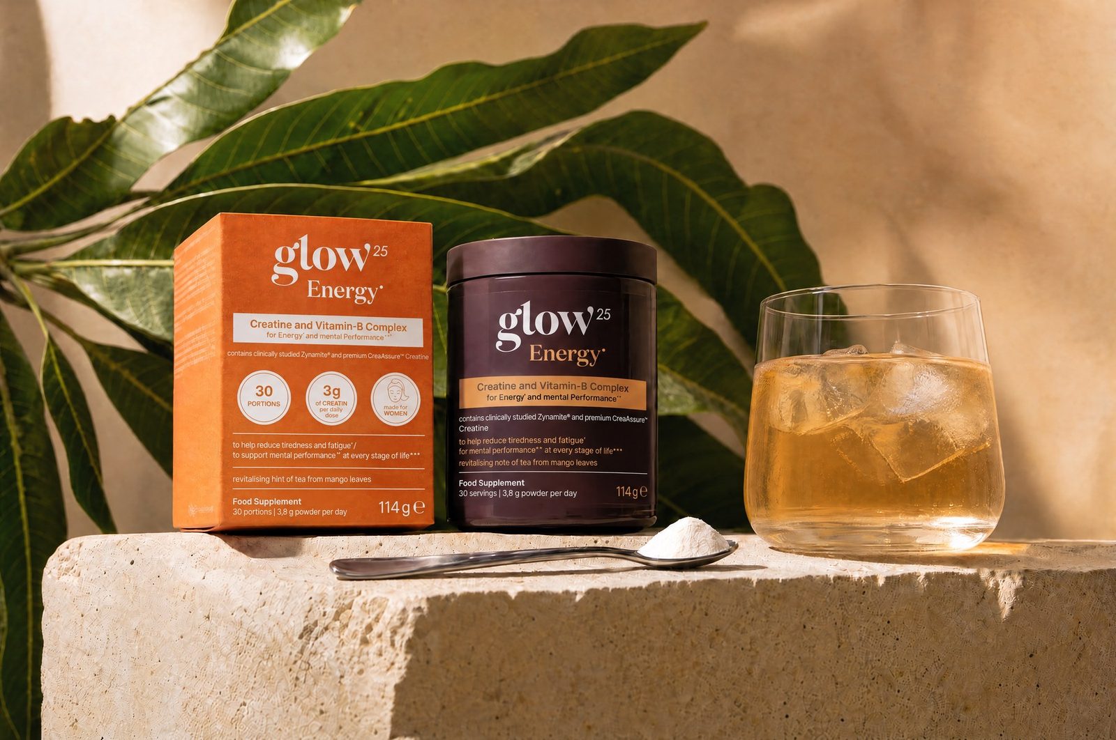 Boîte et pot Glow25 Energy® sur du travertin, à côté des feuilles de manguier, une cuillère et un verre de boisson glacée